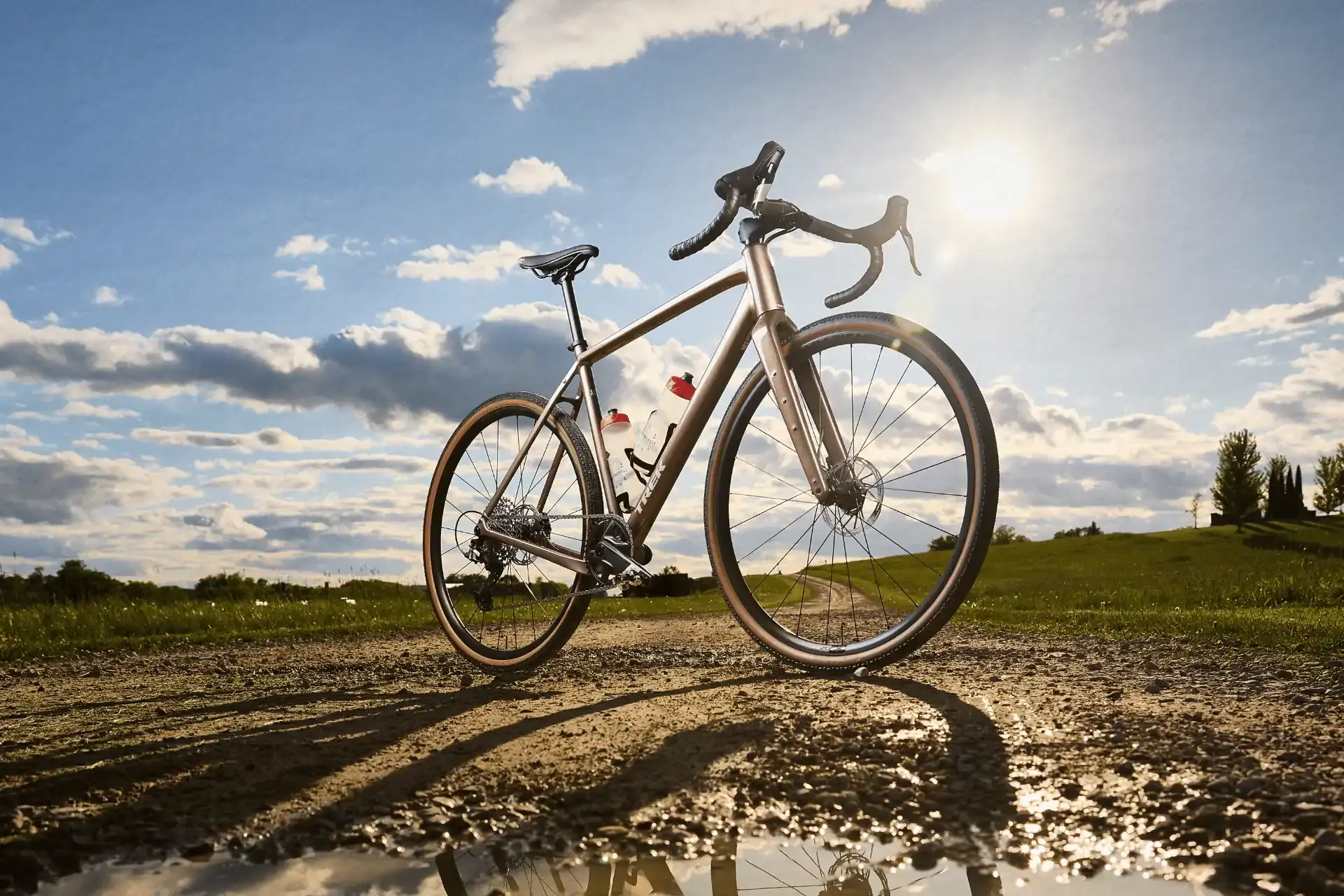 Ein goldfarbenes Gravelbike der Marke Trek steht auf einem schottrigen Pfad im Grasland, während die Sonne im blauen Himmel scheint.