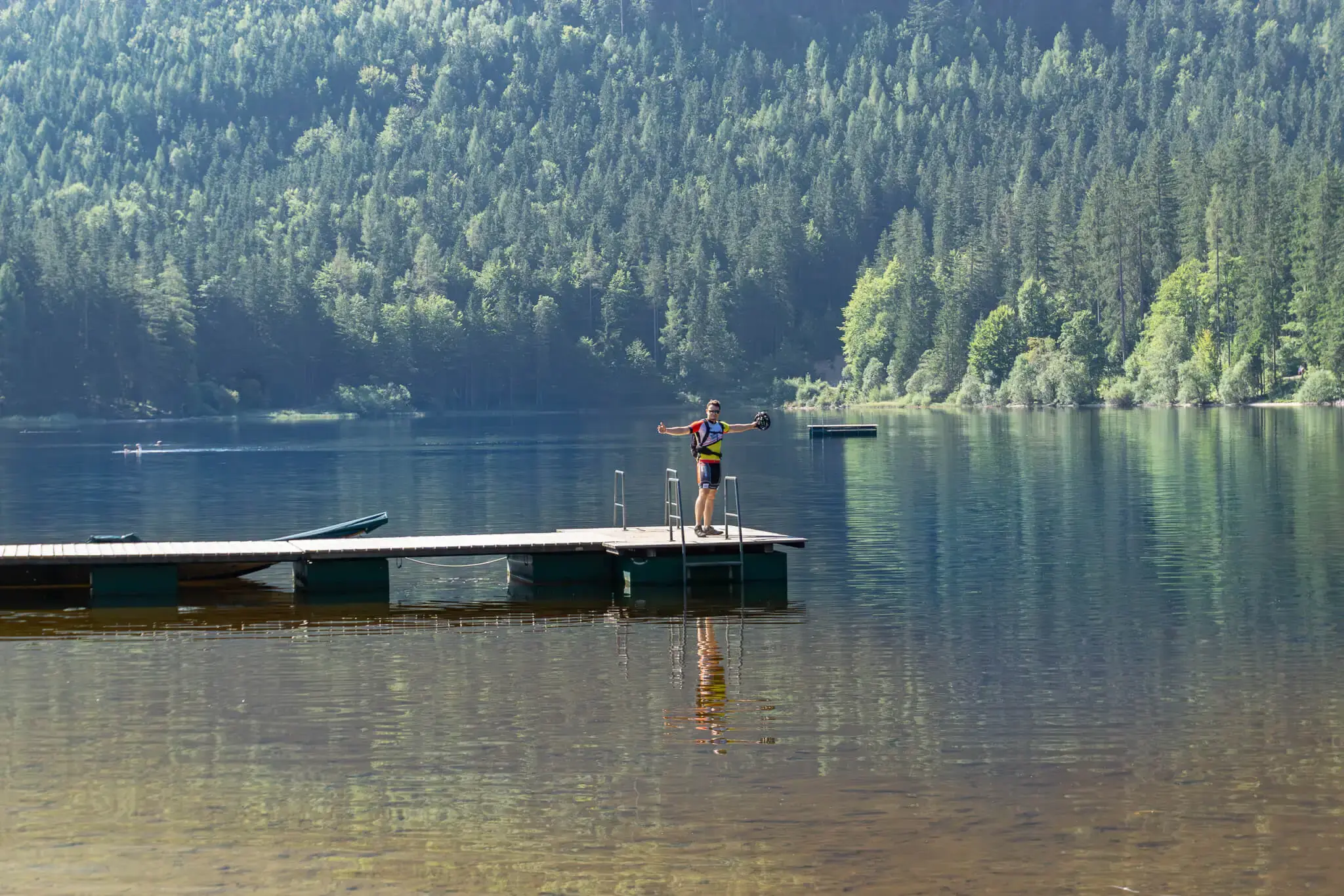 Radfahrer steht in Fahrradkleidung auf einem Holzsteg an einem See.