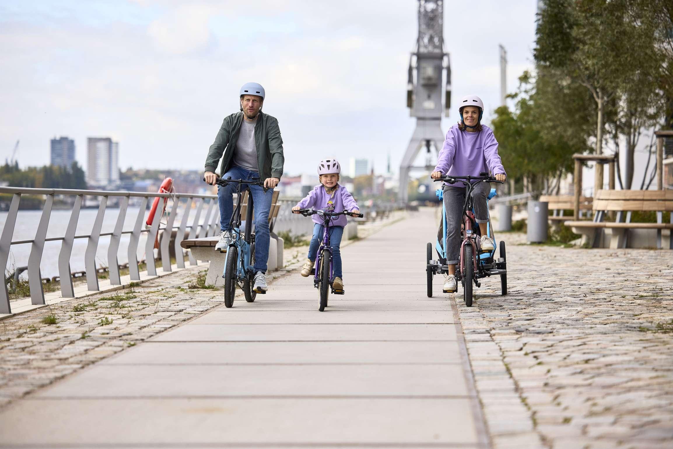 Familie faehrt gemeinsam auf Fahrraedern an einem Flussufer entlang.