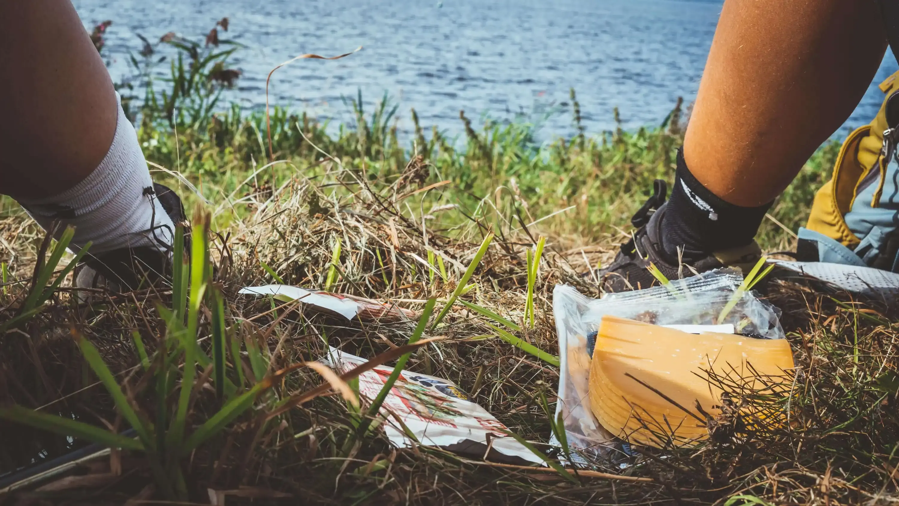 Detailbild: 2 Radfahrer am Fluss, man sieht nur die Beine und etwas zu essen. Im Hintergrund der Fluss.