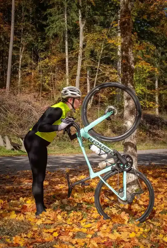 Eine Frau in sportlicher Kleidung lehnt ihr hellblaues Bianchi-Fahrrad an einem Baumstamm ab. Sie trägt einen Helm und eine gelbe Weste über einem schwarzen Trikot. Der Radfahrer steht auf einem Waldweg, umgeben von vielen gelben Herbstlaubblättern.