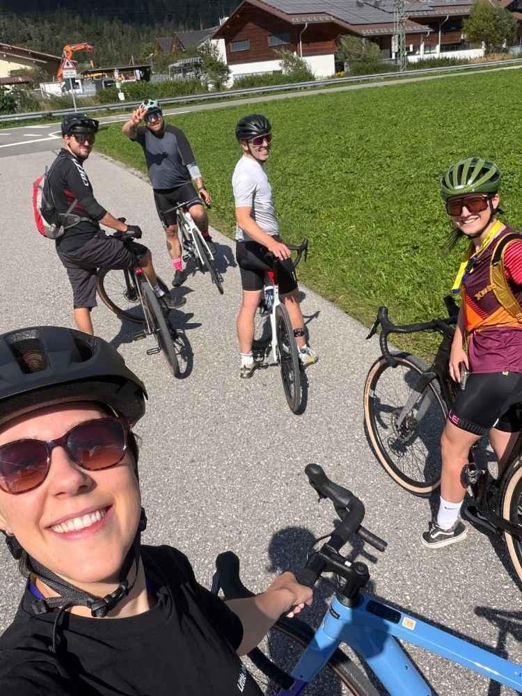 Fünf radfahrende ljB Kollegen mit Helmen stehen mit ihren Fahrrädern auf einer Straße neben einer grünen Wiese