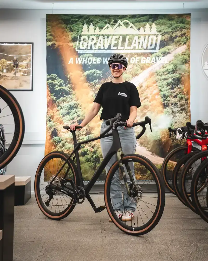 Eine Frau in Helm und Sonnenbrille steht lächelnd in einem Fahrradgeschäft neben einem schwarzen Gravelbike. Im Hintergrund hängt ein großes Wandbanner mit dem Logo Gravelland.