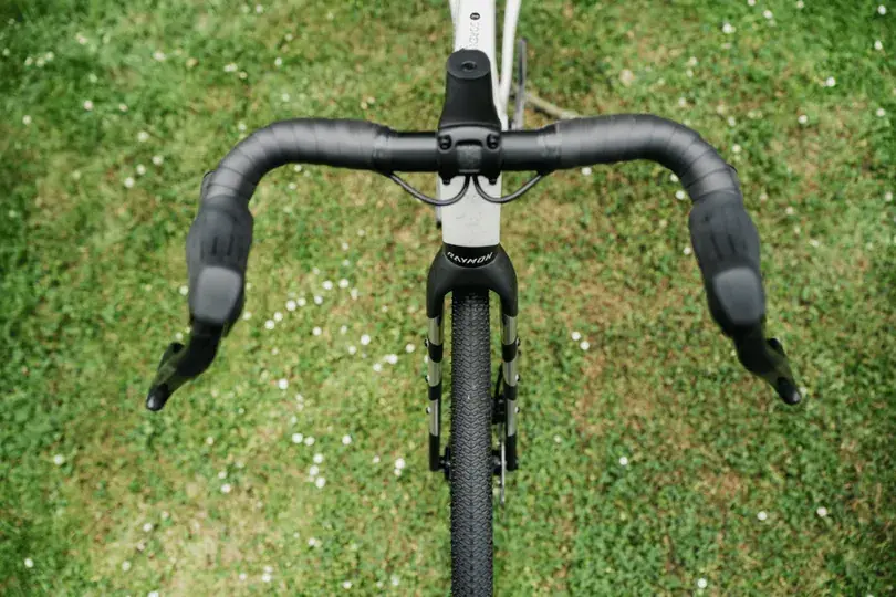 Ein Blick auf die schwarzen Blade Lenker eines weißen Gravelbikes mit der Marke Raymon, das auf grünem Rasen steht.