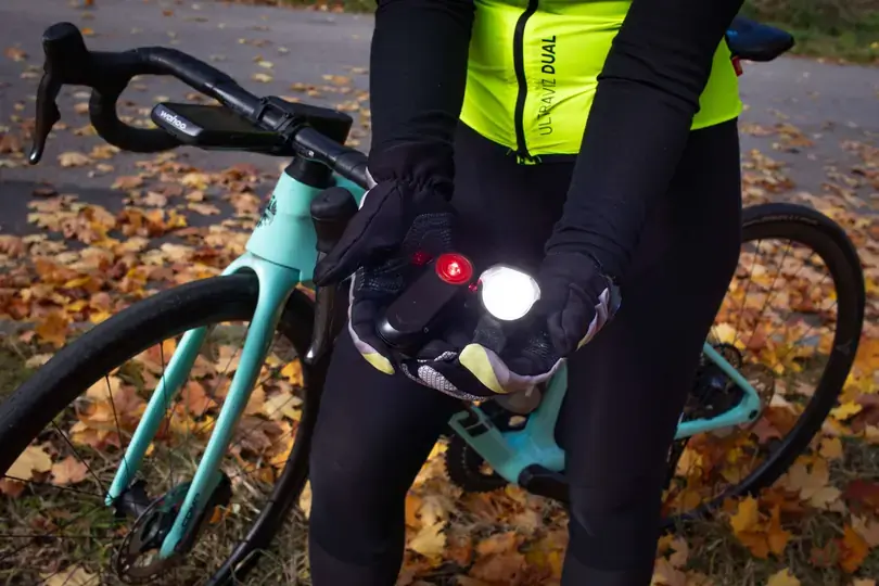 Radfahrer hält in den Händen ein leuchtendes Fahrradlicht und ein Reflektor, während er neben einem hellblauen Rennrad steht, umgeben von Herbstlaub.