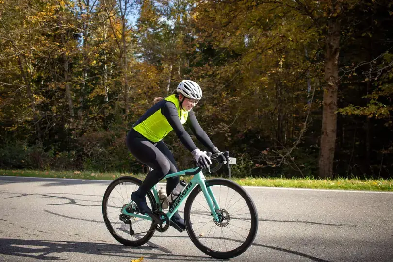 Eine fahrradfahrende Frau in gelber Weste und Helm fährt auf einem hellgrünen Bianchi-Roadbike eine asphaltierte Straße entlang. Der Hintergrund zeigt einen dichten Wald mit gelb gefärbtem Herbstlaub unter blauem Himmel.