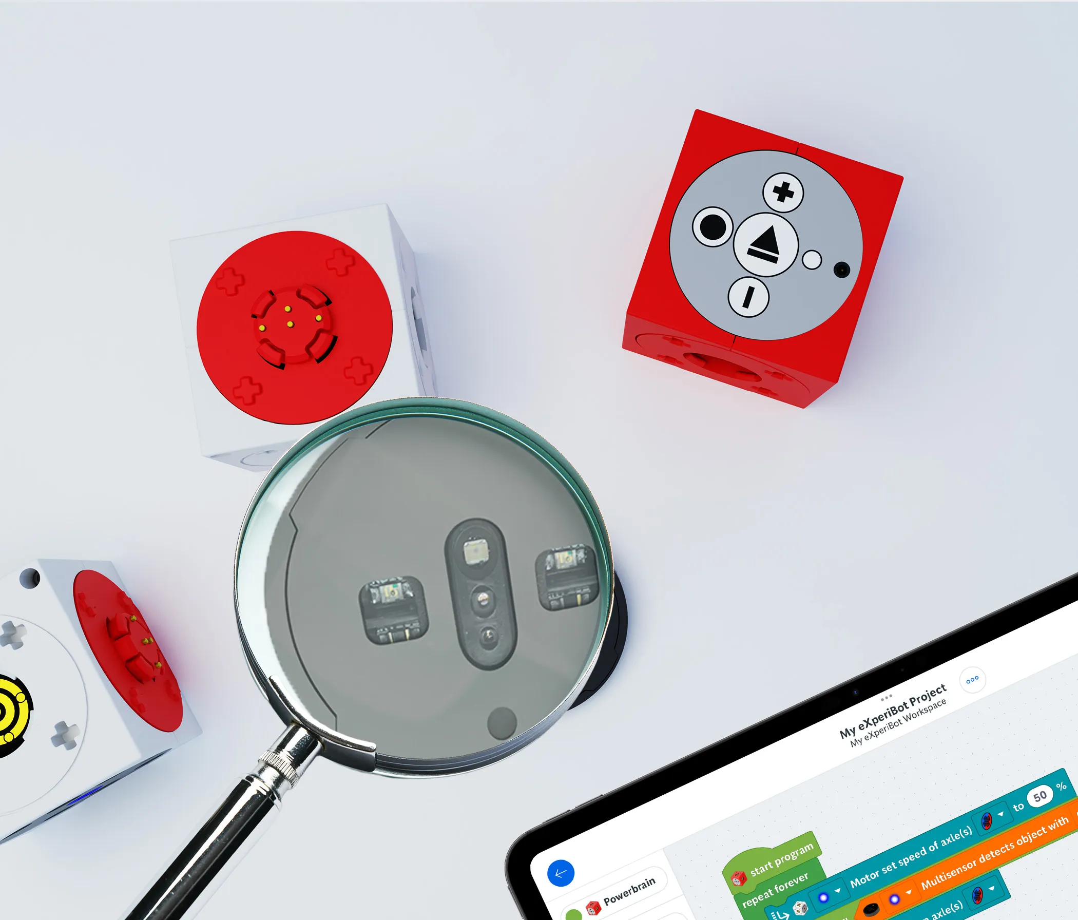 Eine Auswahl an eXperiBot-Modulen und ein Tablet mit der eXperiBot Blockly App. Über den Multisensor wird eine Lupe gehalten.