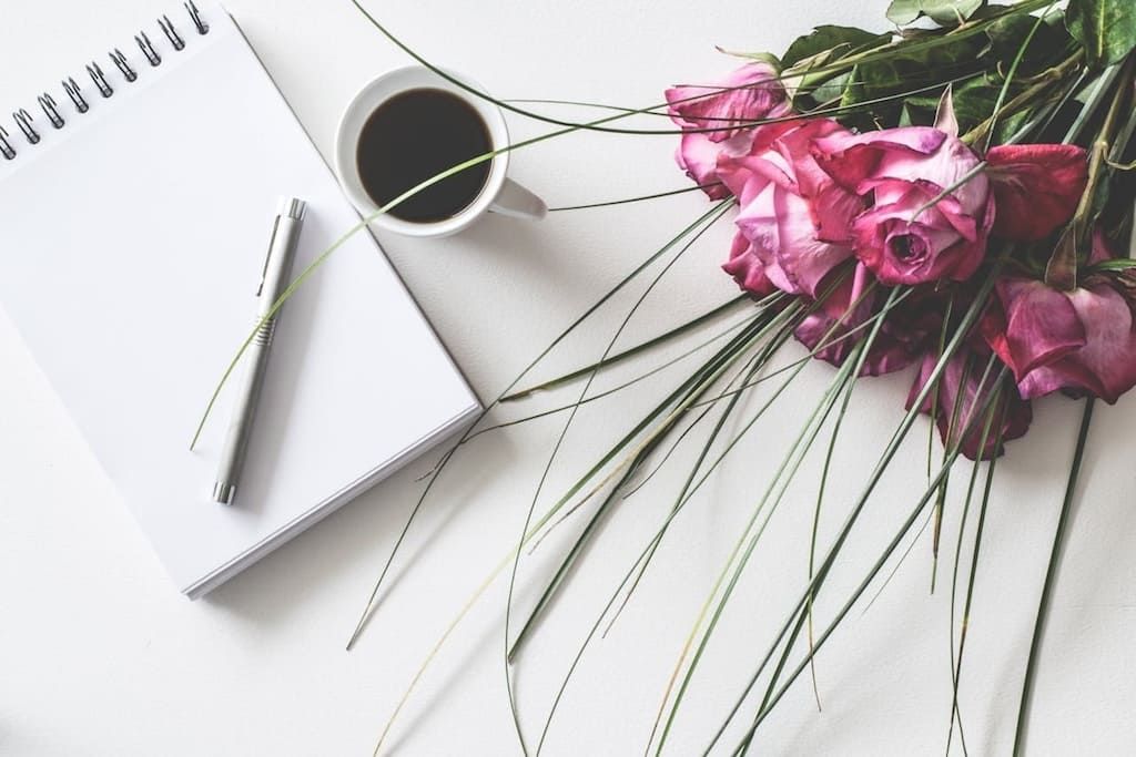 Open notebook with a pen, cup of coffee, and pink flowers on a light desk.