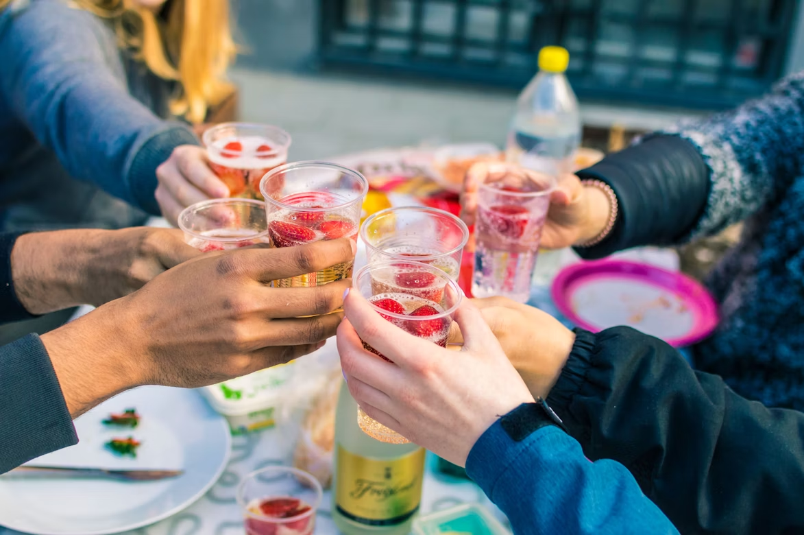 Several people raising plastic cups filled with sparkling drinks and strawberries for a toast at an outdoor gathering.
