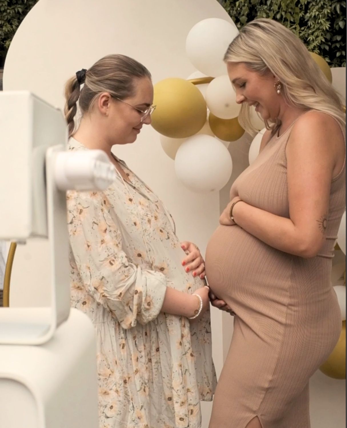 Two smiling pregnant women stand facing each other, looking down. They are both holding their baby bumps. The woman on the left wears a light-colored, floral dress and round glasses, her hair in a high braided ponytail. The woman on the right wears a fitted, beige ribbed sleeveless dress. The background features a white round backdrop arch decorated with clusters of white and gold-colored balloons. In the left corner of the photo, a part of a white photo booth is visible in the foreground.