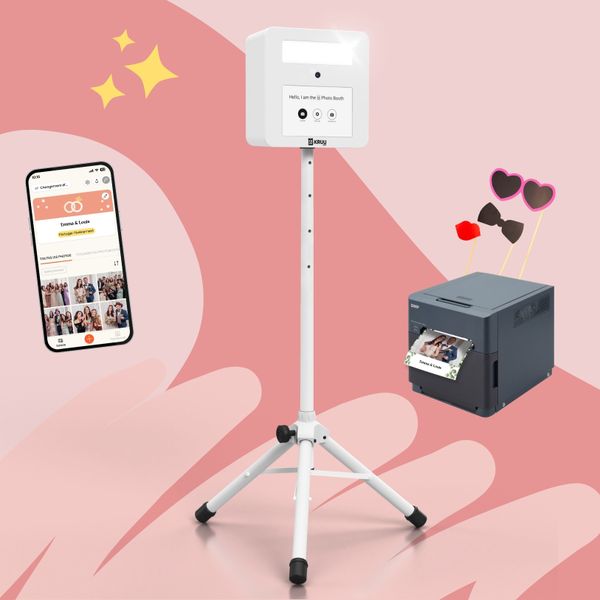 A product layout on a pink background featuring a white KRUU photo booth on a tripod stand in the center. To the left, there is a smartphone displaying a digital event gallery within the KRUU App. To the right, a dark grey DNP printer is shown with a freshly printed photo of a couple. Above the printer, three photo props like heart-shaped glasses, a bow tie and a kissing mouth are assembled.