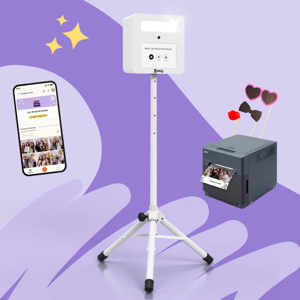 A product layout on a purple background featuring a white KRUU photo booth on a tripod stand. To the left, a smartphone displays a birthday-themed event gallery within the KRUU App. To the right, a dark grey DNP printer shows a freshly printed photo of friends at a party. Above the printer, three photo props like heart-shaped glasses, a bow tie and a kissing mouth are assembled.