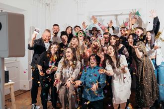 Ein lebhaftes Foto des KRUU-Teams, das vor einem weißen Fotoautomaten auf der linken Seite feiert. Die Mitarbeiter lachen und werfen buntes Konfetti in die Kamera, während sie festliche Accessoires wie übergroße Brillen tragen. Die Szene fängt ein energiegeladenes und fröhliches Gemeinschaftsgefühl ein.