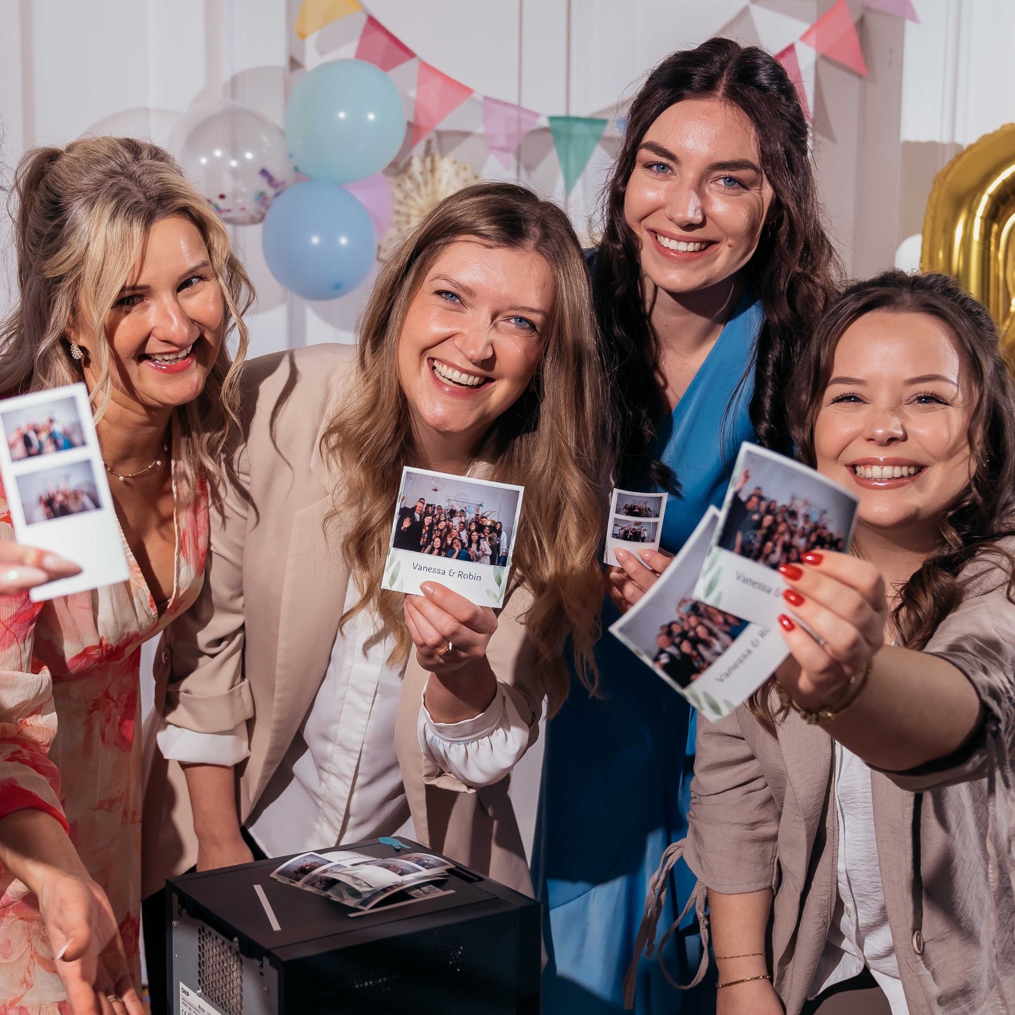 Four smiling women in formal attire are holding up various photo booth prints toward the camera. The prints feature group photos and are personalized with the names "Vanessa & Robin." In the foreground, a photo printer is visible with more photo strips resting on top. The background is festively decorated with light blue and golden foil balloons as well as a colorful pennant banner.