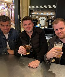 The three KRUU founders Oli, Jochen and Philipp sitting at a bar, each holding a drink, smiling at the camera. Shirts displayed in the background.