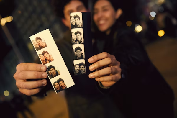 Photo booth fun: Couple holding print strips at night. Happy date memory with instant photos & city lights background.