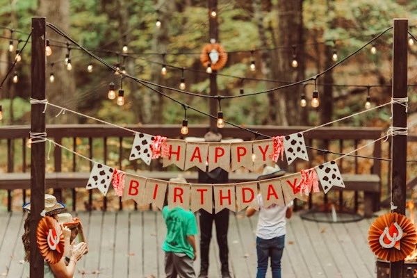 Outdoor birthday setup with string lights and a “Happy Birthday” banner, with people gathered on a wooden deck in the background.