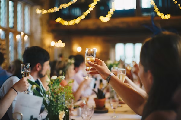 Wedding reception toast: Guest raising champagne glass at dinner. Elegant party celebration with warm string lights.