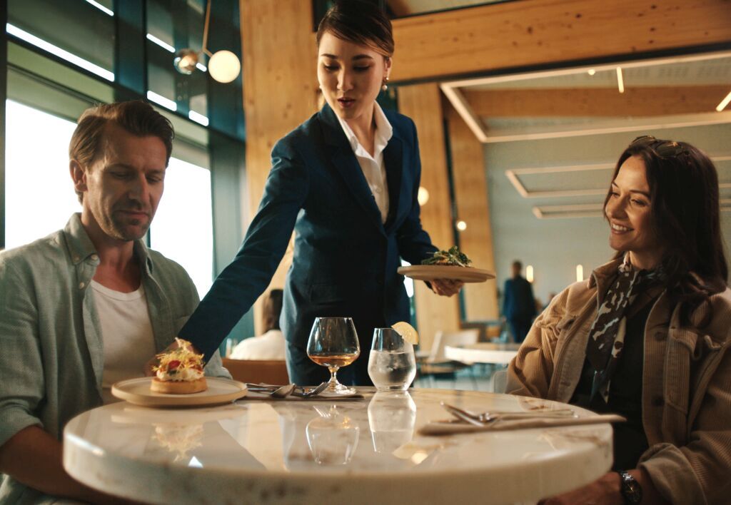 Server in blue uniform serves food to a man and woman at a modern restaurant table set with drinks and utensils.