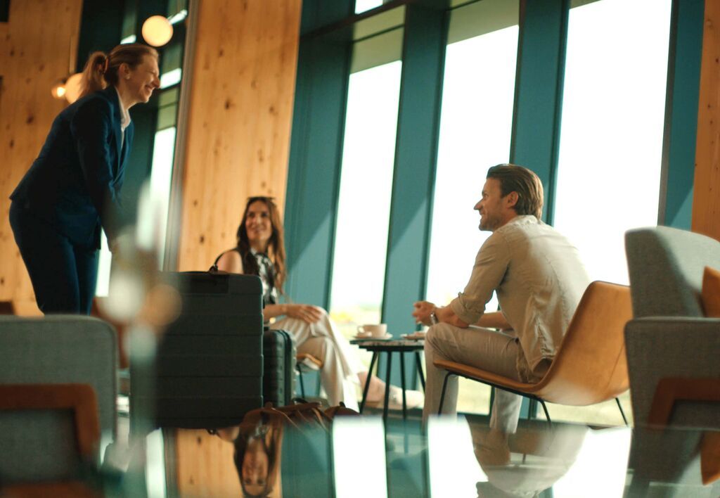 Three people in a modern lounge, engaged in conversation; two sitting, one standing near a large window with natural wood accents around.