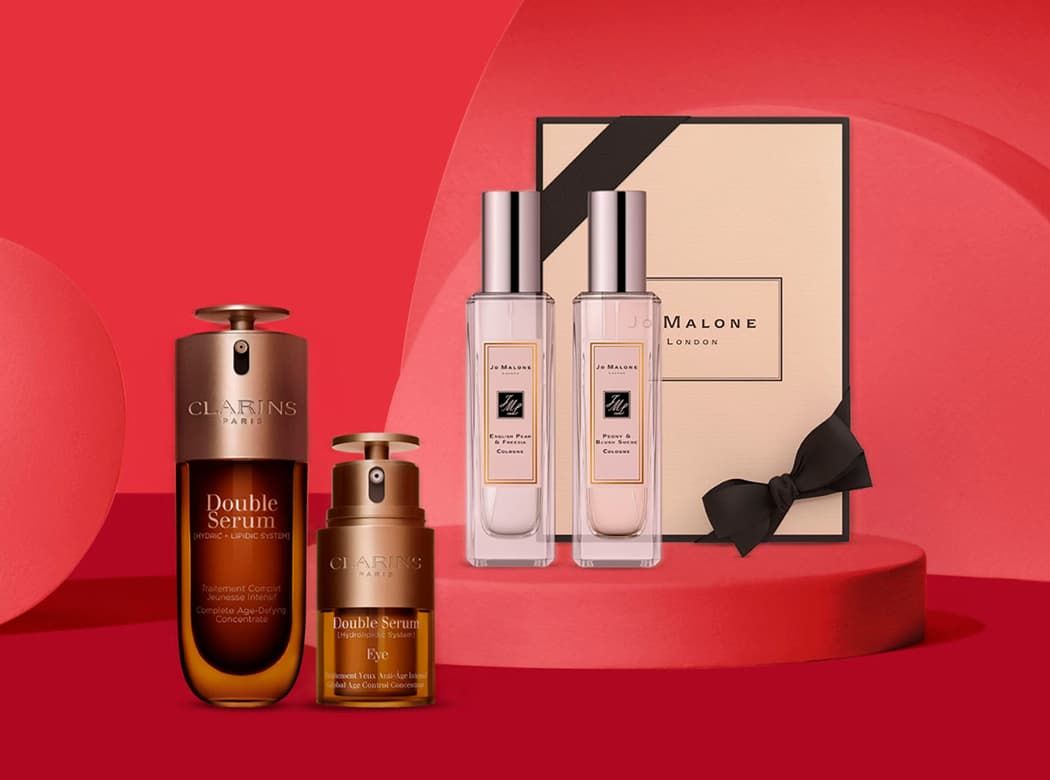 Clarins serums and Jo Malone perfume set against a red background, featuring a gift box and bottles with sleek designs.