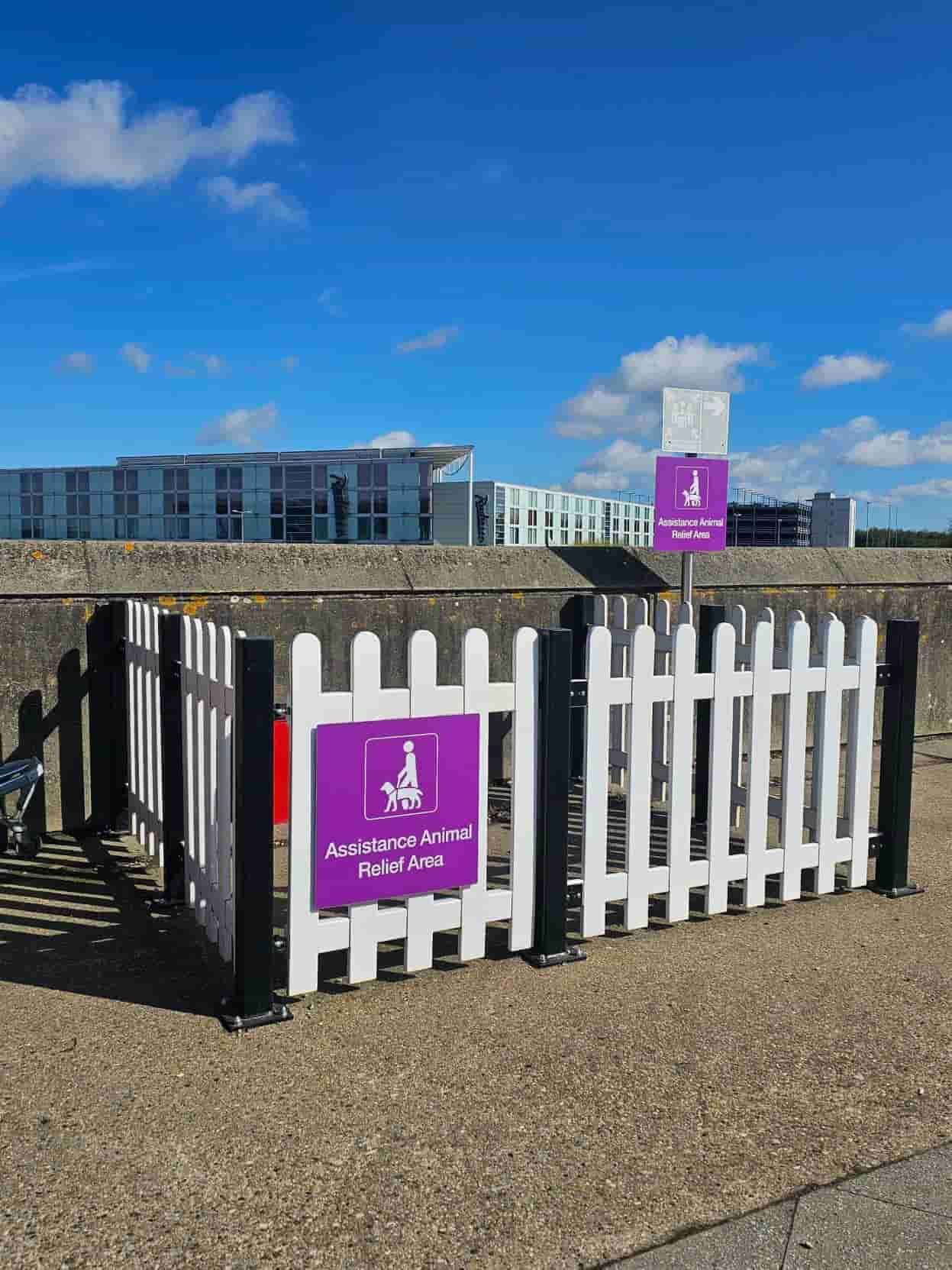 White fenced "Assistance Animal Relief Area" with purple signage, set outdoors under a clear blue sky near modern buildings.