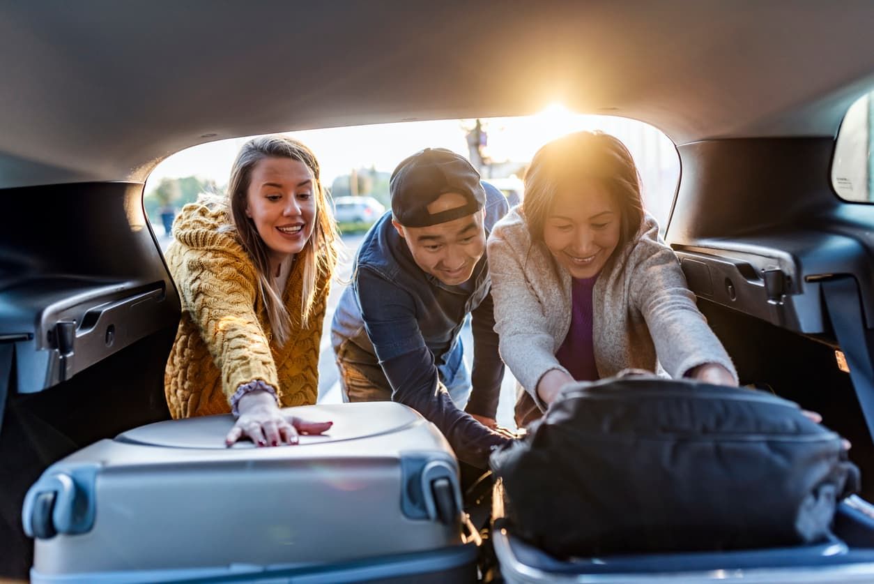 Three smiling people putting luggage in car boot