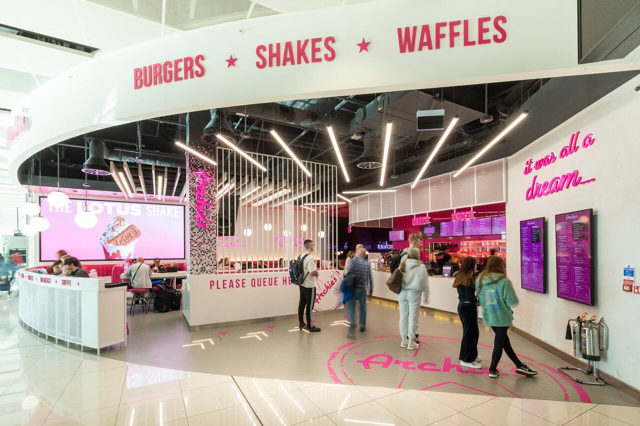 Colorful diner interior with a retro theme, menu signage, and customers queuing. Bright pink accents highlight "Burgers," "Shakes," "Waffles."