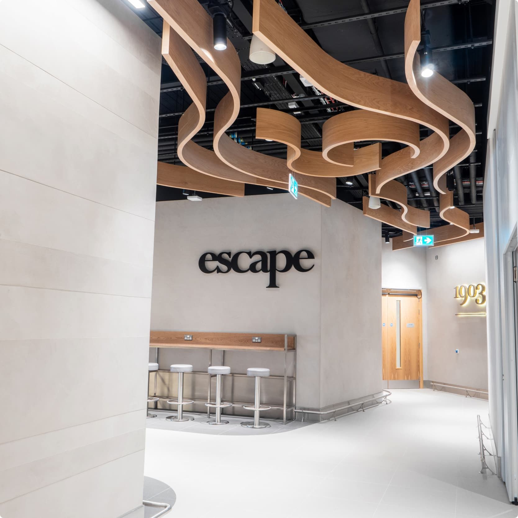 Modern interior with wooden wave ceiling, a sign reading "escape," stools at a counter, and wooden doors on white walls.