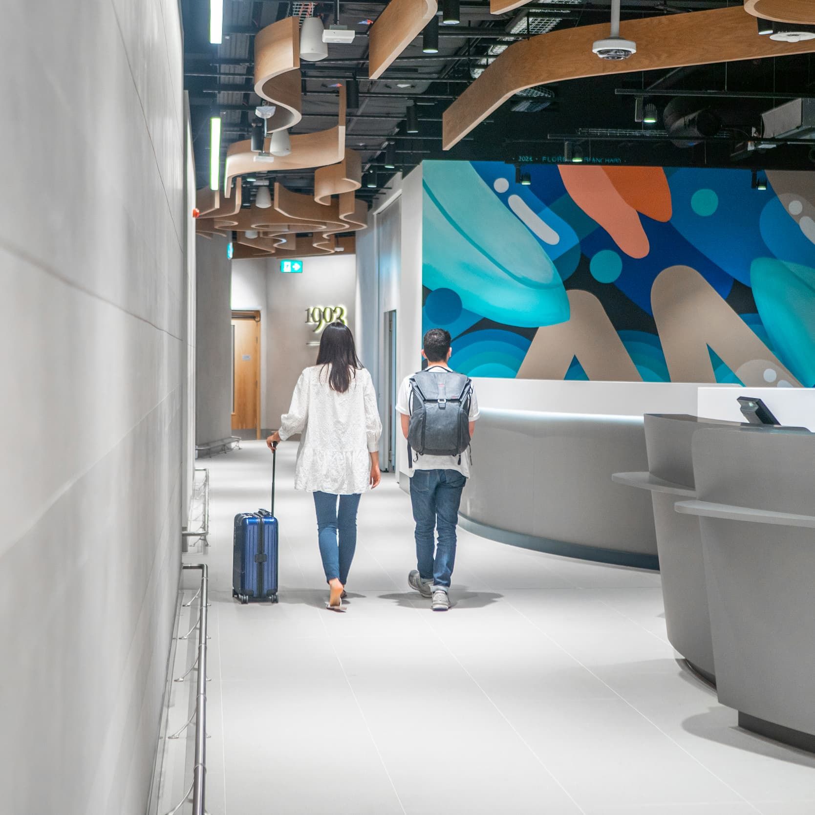 Two people with luggage walking down a modern hallway with vibrant wall art, heading towards a door, past a curved reception desk.