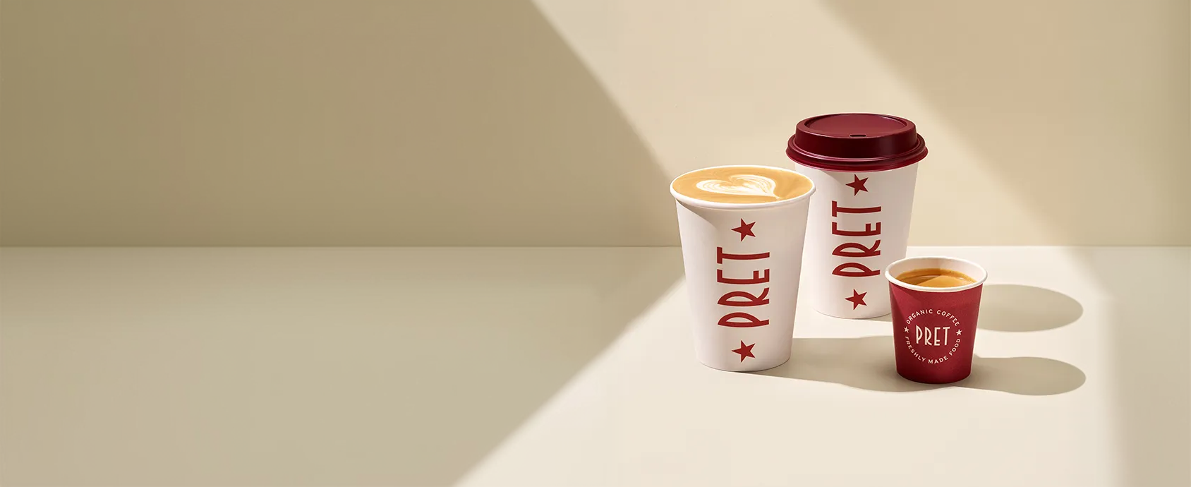 Three Pret coffee cups of varying sizes on a sunlit surface, featuring white and red designs, with two open cups showing frothy coffee.