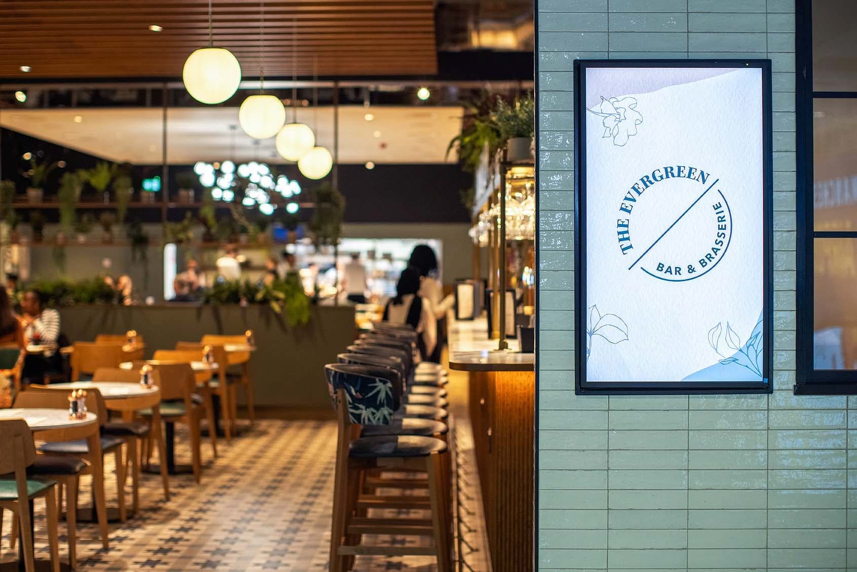 Modern bar and brasserie interior with green tiles, wooden decor, bar stools, and tables. A sign reads "The Evergreen Bar & Brasserie."