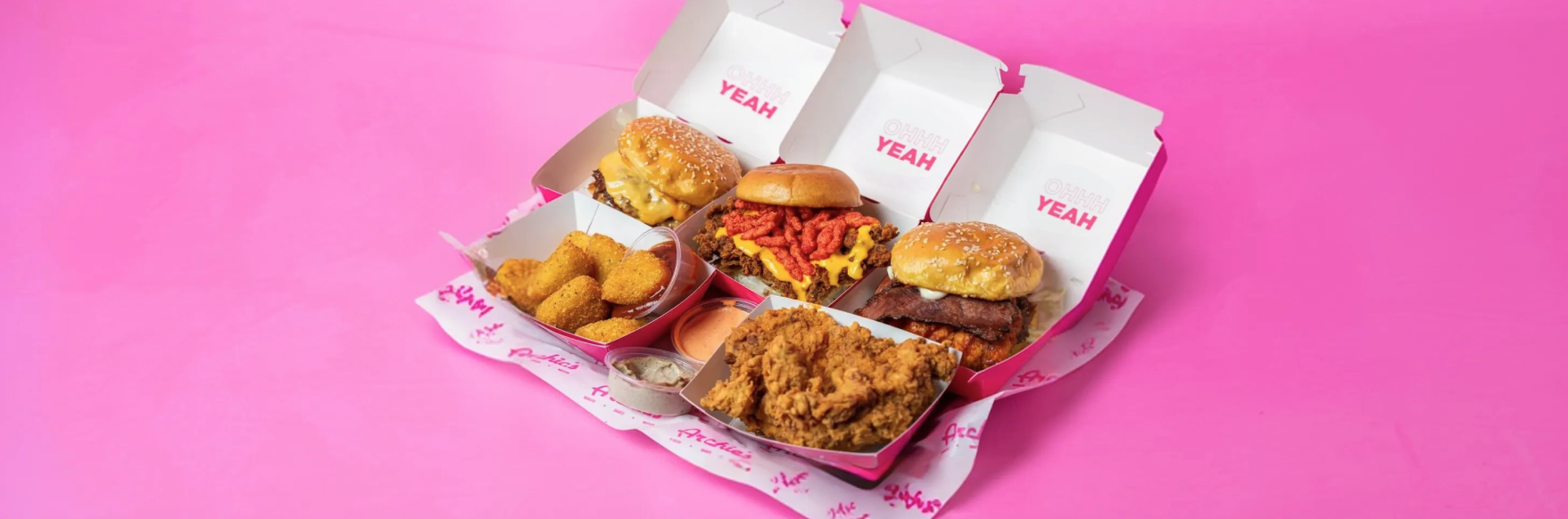 A variety of fast food items on pink packaging: two burgers, fries topped with cheese and chips, nuggets, fried chicken, and dipping sauces.