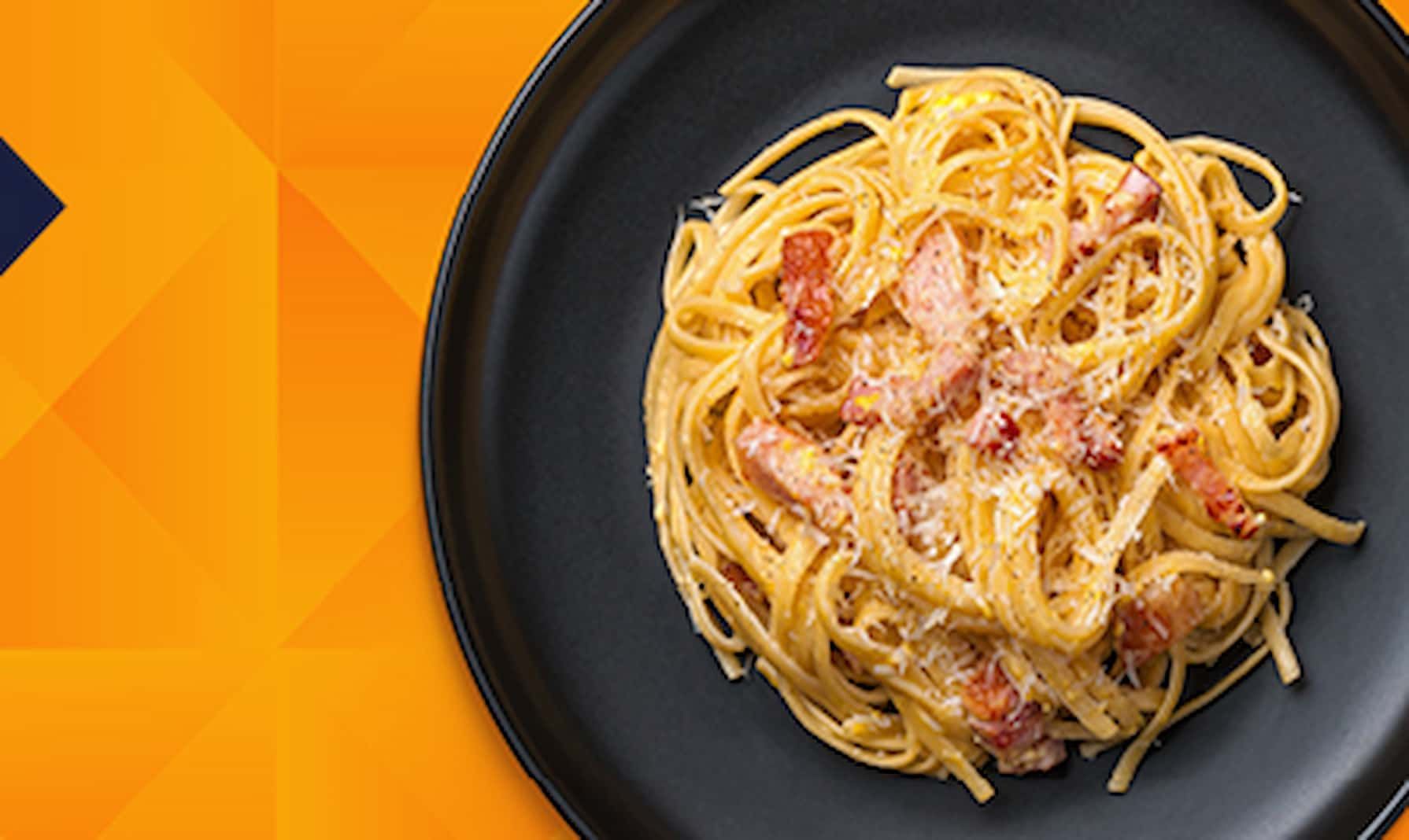 A plate of creamy spaghetti carbonara topped with grated cheese and bacon, served on a black plate against an orange geometric background.