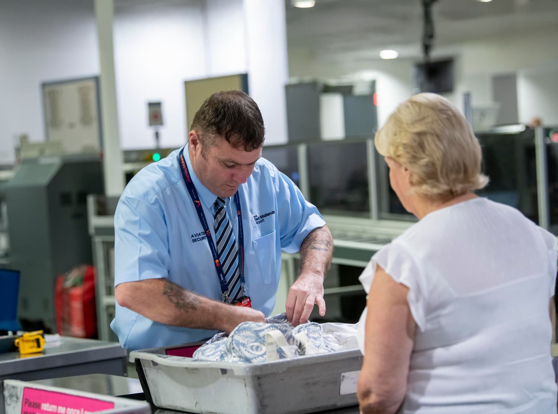 Airport security personnel inspects a passenger's belongings in a tray at a screening area, with large equipment visible in the background.