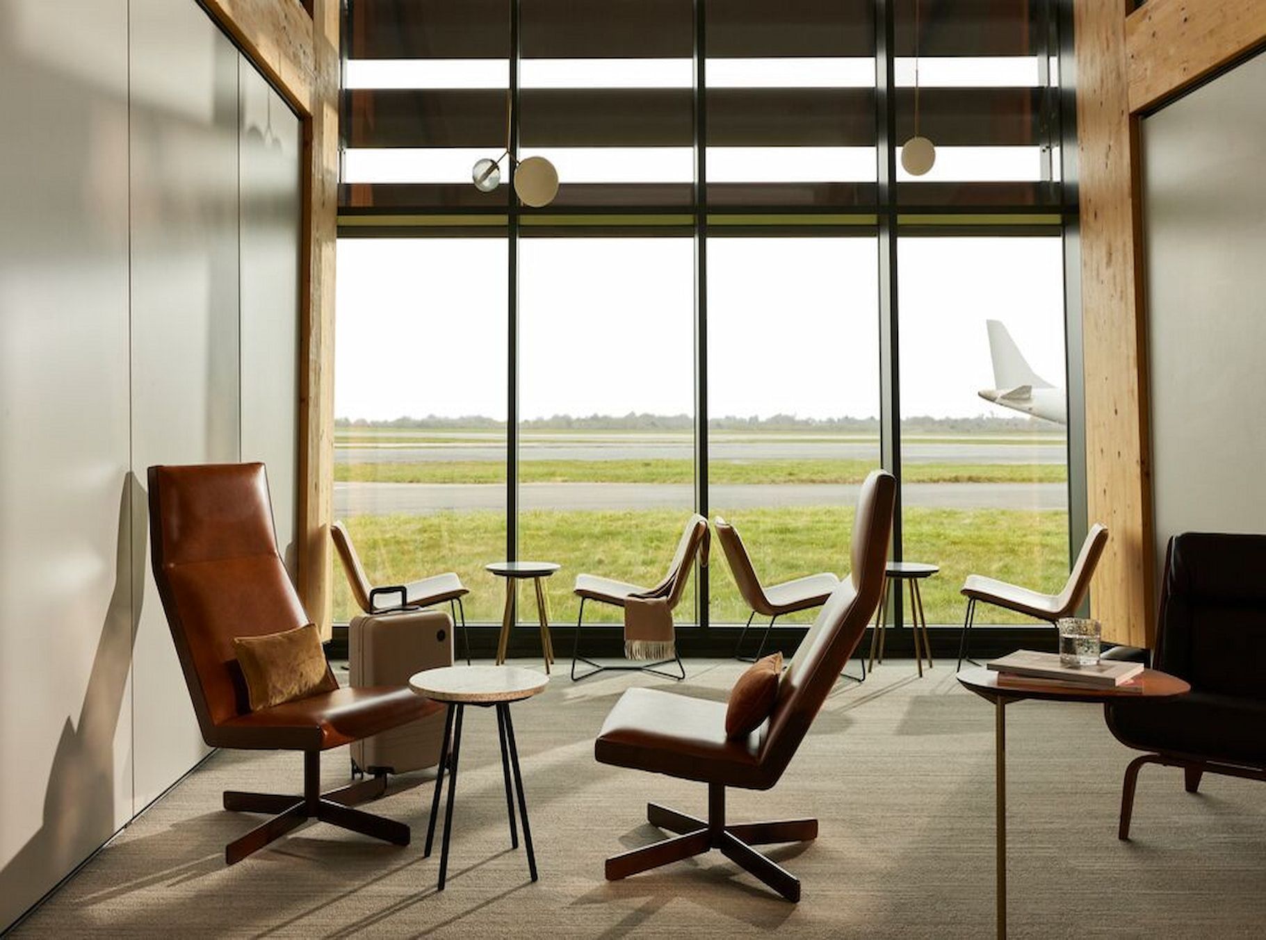 Modern airport lounge with leather chairs and tables, large windows overlooking a runway with a plane tail visible outside.