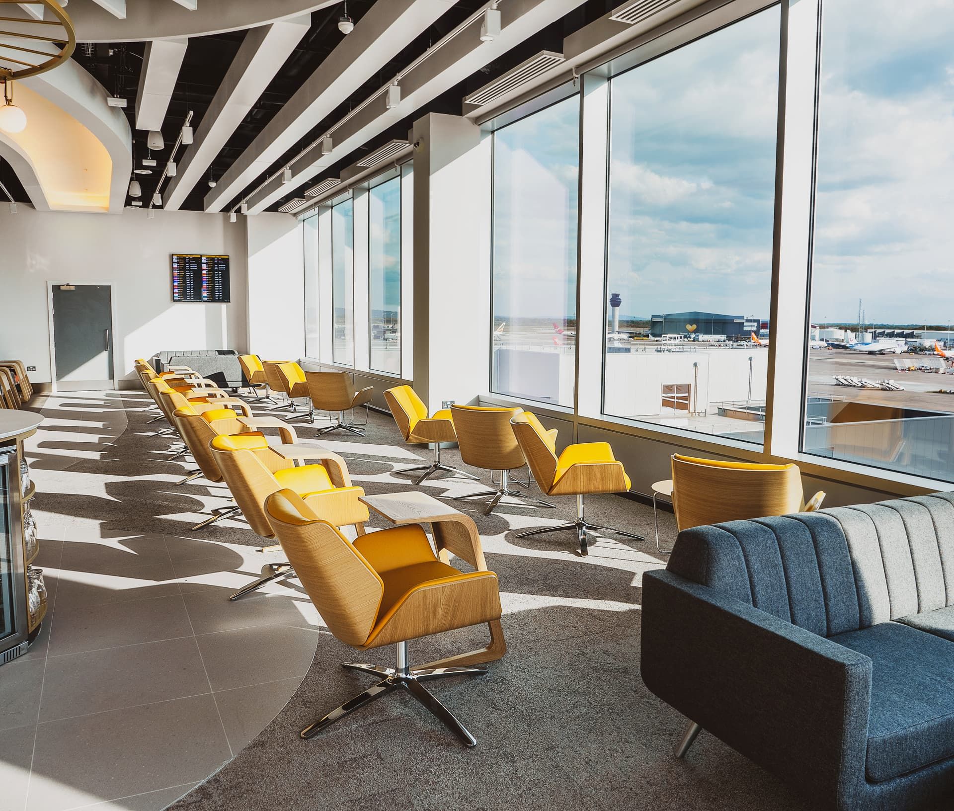 Modern airport lounge with yellow chairs, large windows viewing the runway, a dark gray sofa, and a flight information screen on the wall.