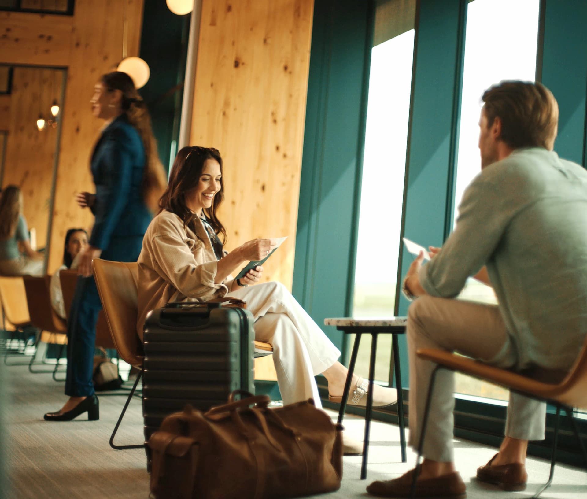 Woman sitting with luggage, smiling and holding a ticket in an airport lounge, engaging with a man in casual attire.