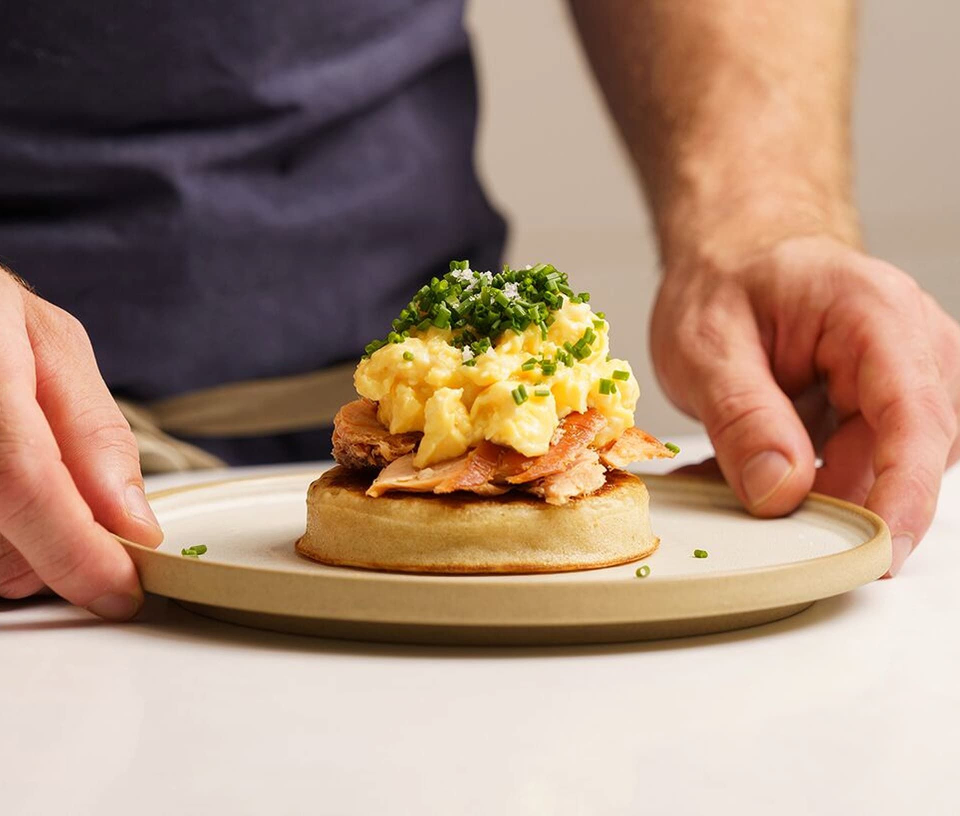 Hands holding a plate with a dish featuring scrambled eggs, bacon, and chopped chives on a round bread base.