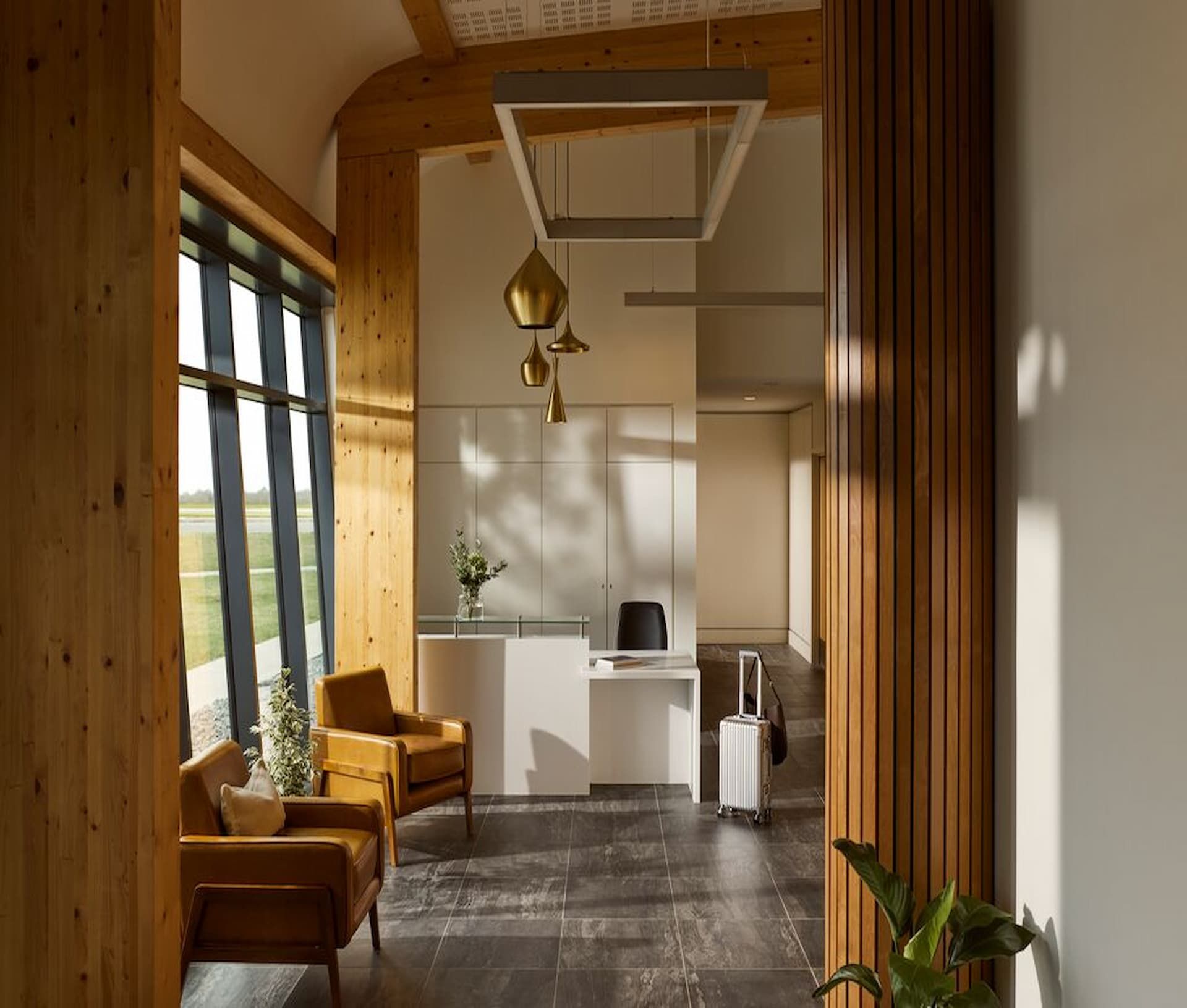 Modern office lobby with wooden accents, large windows, warm lighting, leather chairs, a sleek white desk, and a suitcase near the entrance.