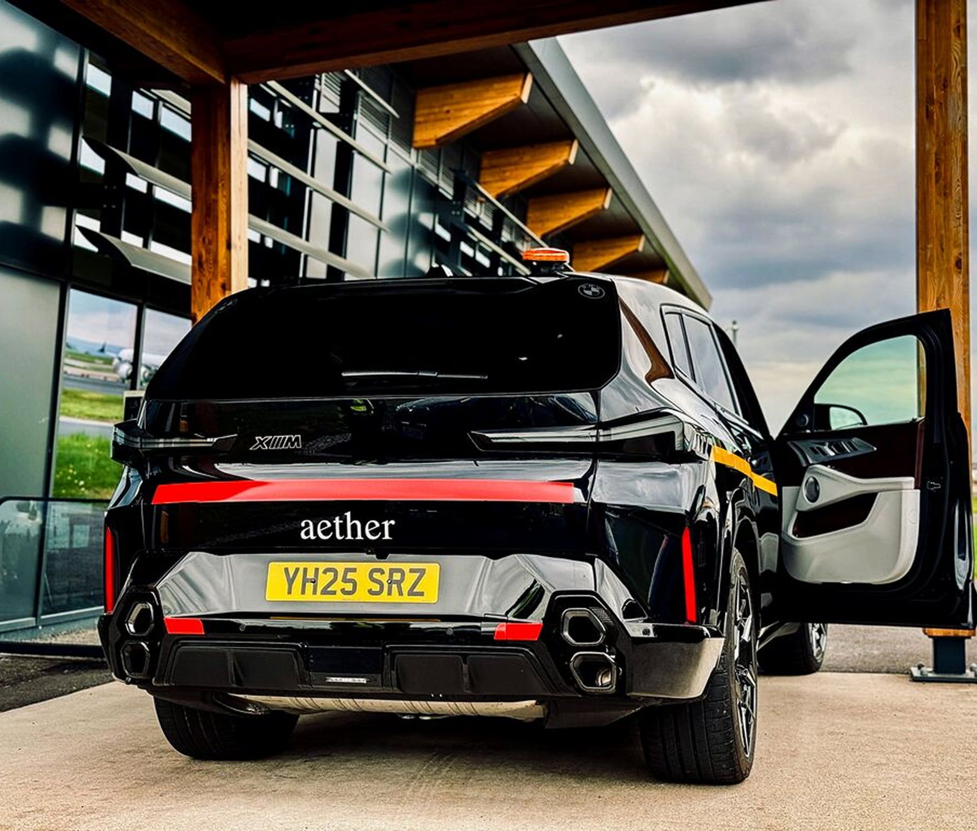 Black SUV with tinted windows, UK license plate YH25 SRZ. Parked by a modern building, one door open, under a cloudy sky.