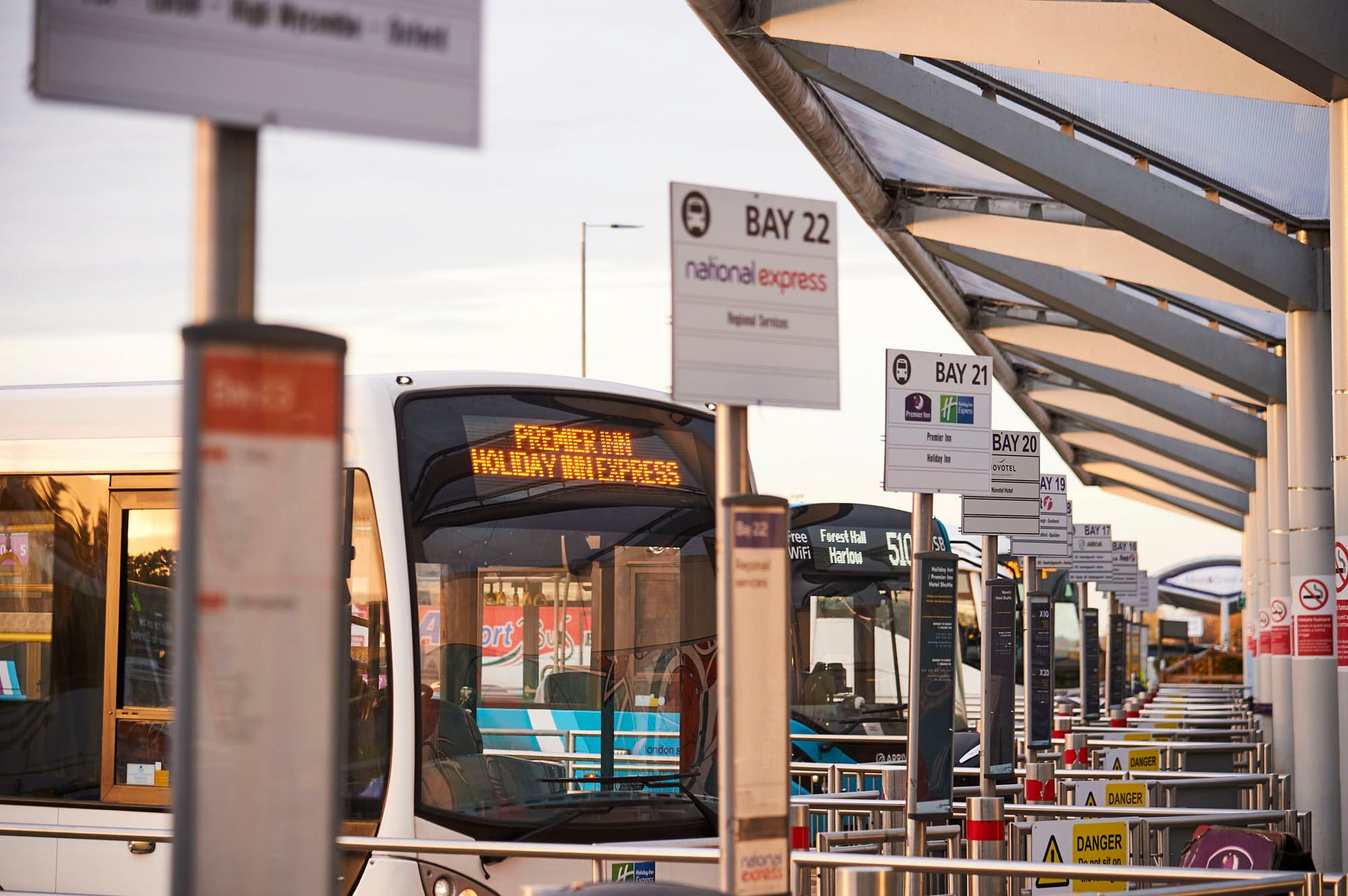Bus terminal with multiple buses parked at bays, including Bay 22. Signs display destinations and warnings. Passengers are visible boarding.