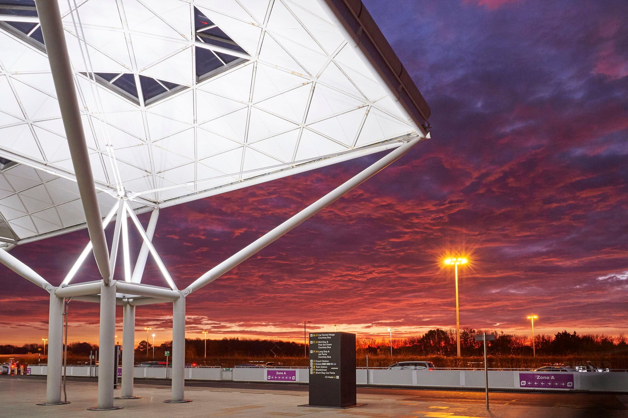 Modern airport terminal with illuminated geometric roof against a vibrant sunset sky, featuring deep purple and orange hues.