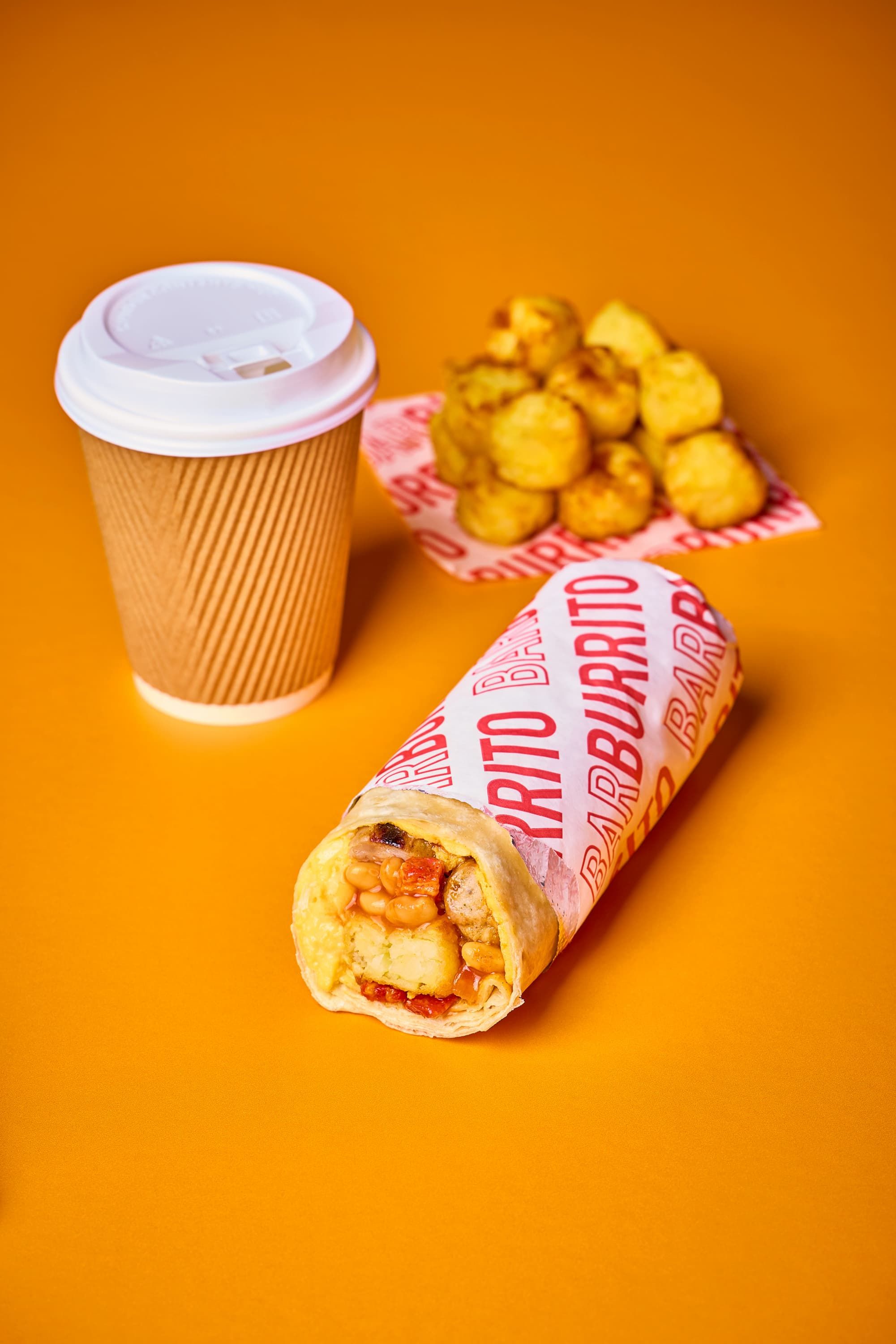 Burrito wrapped in branded paper with a side of tater tots on a pink tray, accompanied by a disposable coffee cup, set on an orange surface.