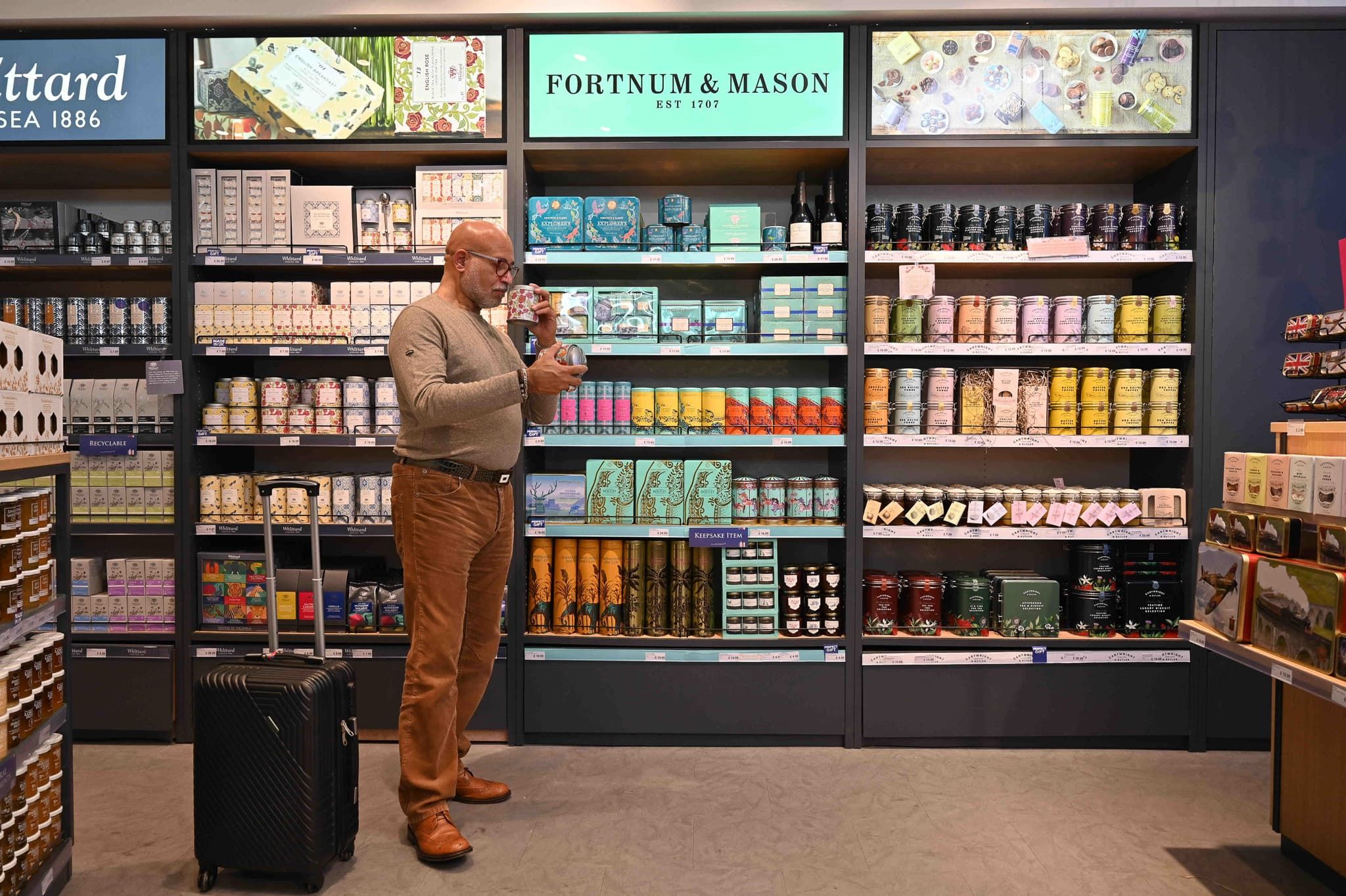 A man with a rolling suitcase examines a tea tin in a Fortnum & Mason store aisle lined with colorful tea and gift tins.