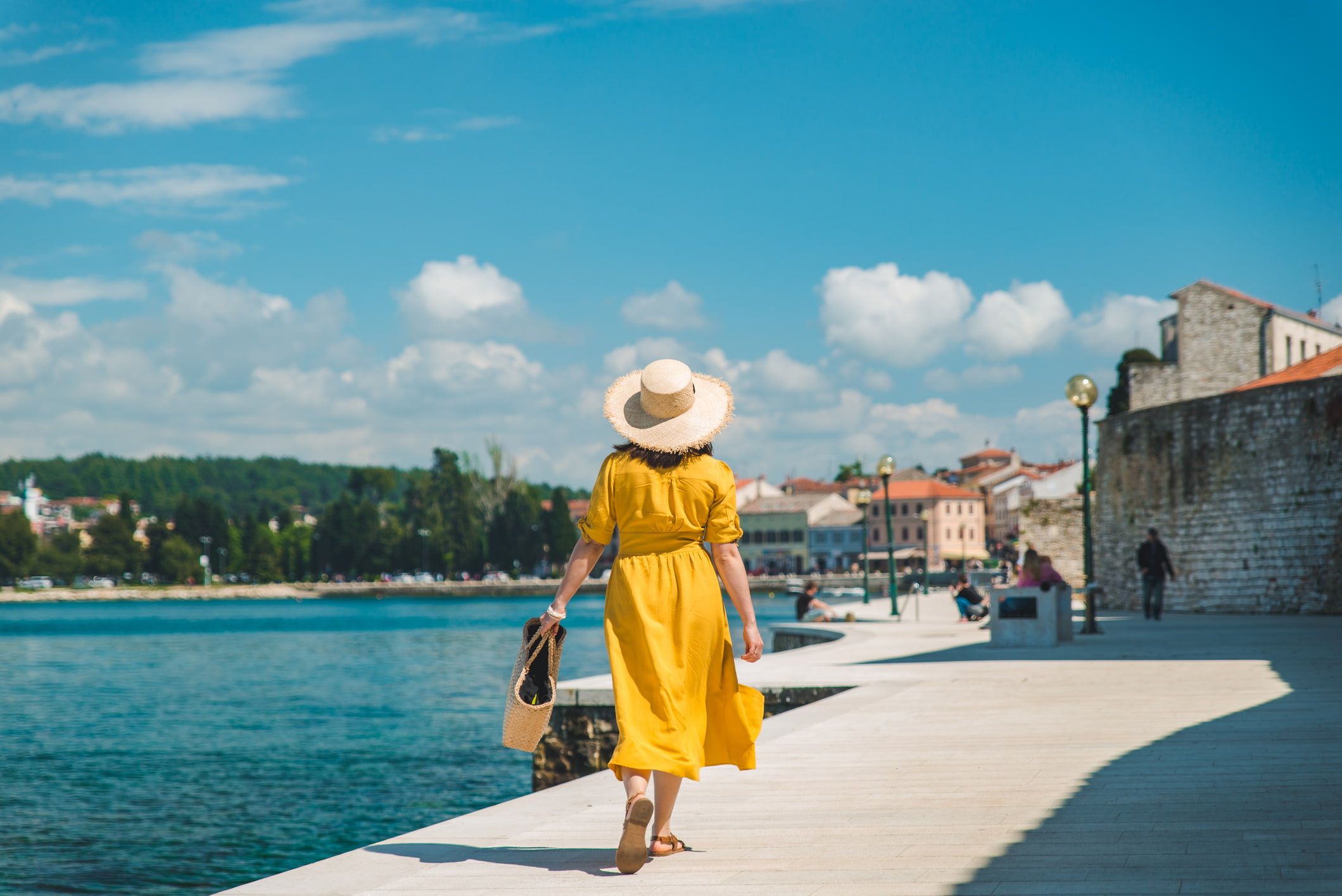 A woman in a yellow dress and straw hat walks along a sunlit waterfront promenade, holding a woven bag, with a town and trees in the background.