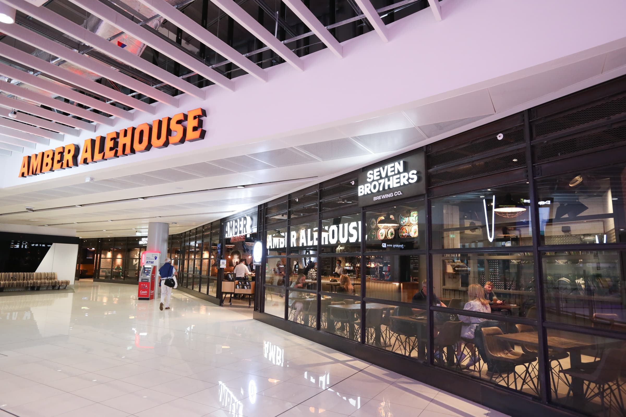 Modern airport restaurant with glass facade, "AMBER ALEHOUSE" and "SEVEN BROTHERS" signs, and people dining inside. Bright, spacious interior.