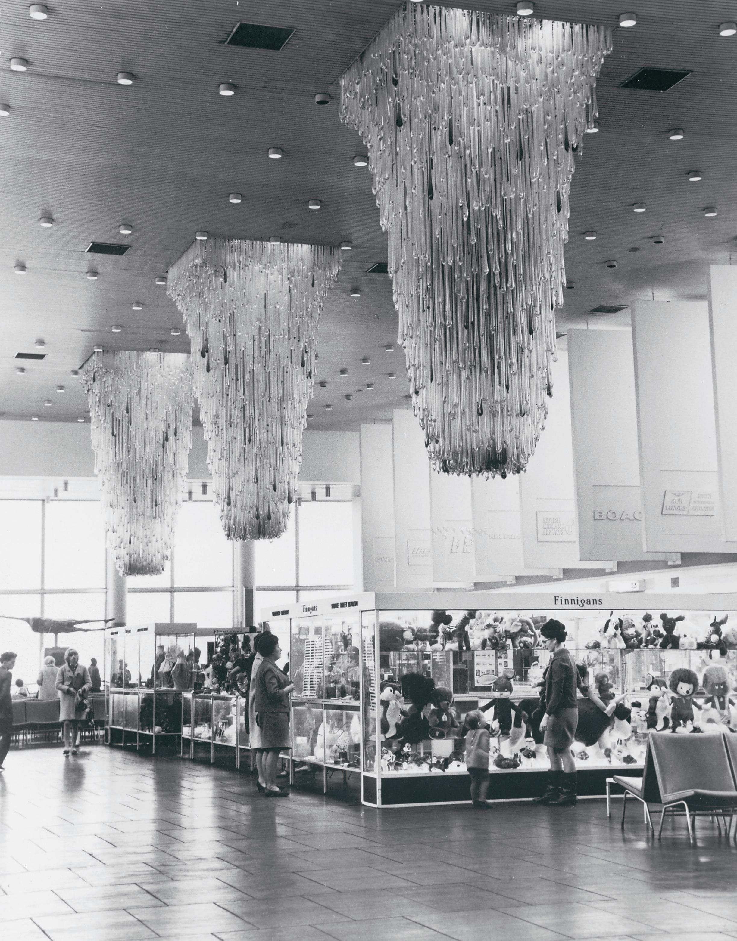 People browse display cases in a large room with elaborate chandeliers hanging from the ceiling, creating a striking pattern above.