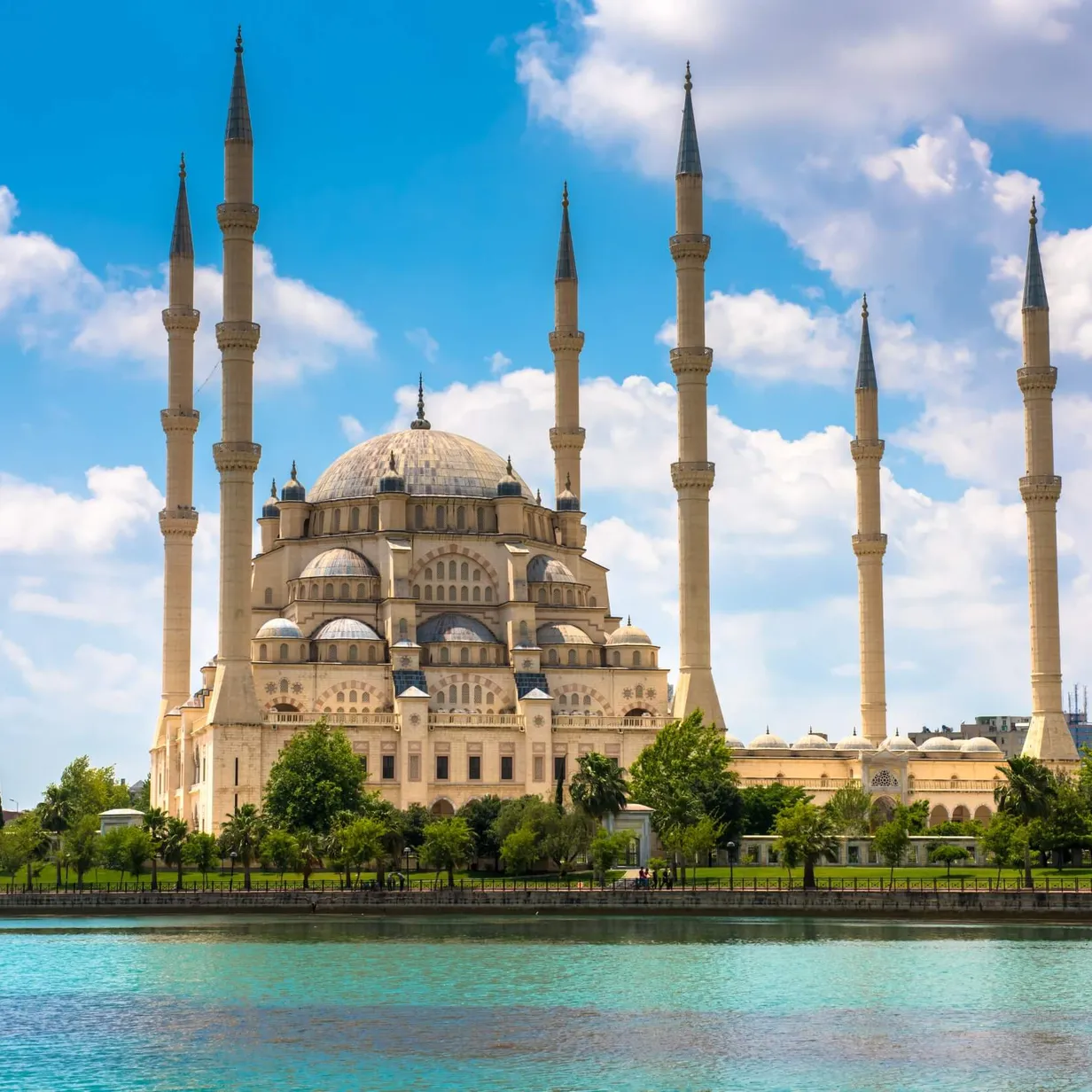 Image of Adana Ulu Camii, the great mosque. The mosque is a large white struture with multiple spires, set amongst lush green gardens and surrounded by water.