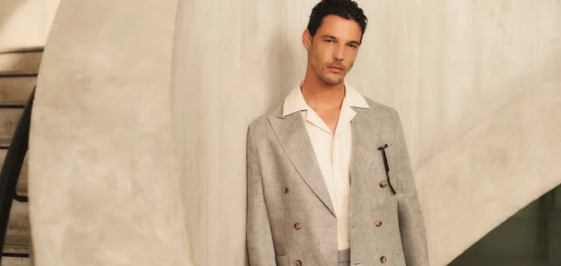 Man in a light grey suit leans against a curved concrete wall, gazing forward with a calm expression.